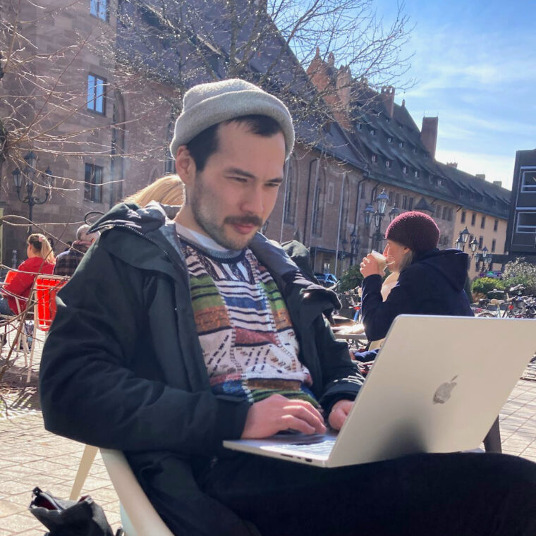 Jeff is sitting outside a café in good weather with a MacBook on his lap.