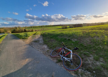 Ein rotes Rennrad liegt am Straßenrand vor einer hügeligen Abfahrt bei schönem, blauem Himmel und sich anbahnendem Sonnenuntergang.